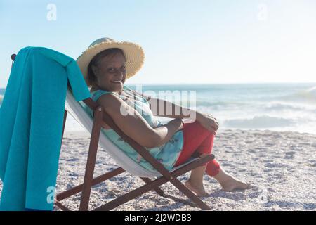 Ritratto di sorridente donna anziana afroamericana che indossa un cappello seduto su una sedia pieghevole in spiaggia Foto Stock