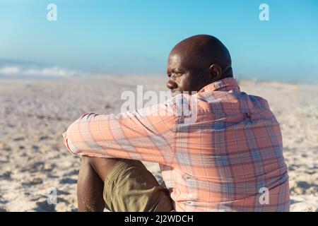 Vista posteriore di un anziano afro-americano in pensione che guarda via mentre si siede in spiaggia in giorno di sole Foto Stock