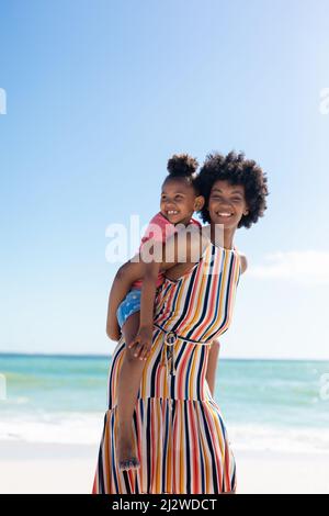 Ritratto di felice madre afroamericana che dà giro di piggyback alla figlia alla spiaggia contro il cielo Foto Stock