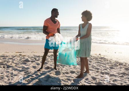 Felice coppia afroamericana raccolta rifiuti insieme in sacchetto di plastica in spiaggia il giorno di sole Foto Stock