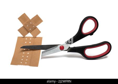 Primo piano delle forbici tagliando un grande pezzo di medicazione per il trattamento di piccole ferite sulla pelle sotto due cerotti incrociati bruni attaccanti contro bianco b Foto Stock