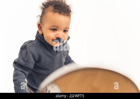 Adorabile ragazzo divertente biraciale con baffi neri finti che si guarda nello specchio su sfondo bianco. Foto studio. Foto di alta qualità Foto Stock