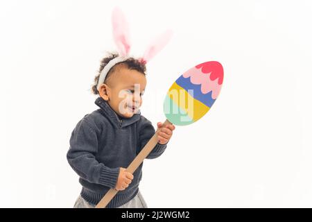 Pasqua e concetto di infanzia. Felice stupito piccolo biraciale bambino ragazzo indossando coniglietto orecchie fascia e tenendo carta uovo di Pasqua posa su sfondo bianco. Foto di alta qualità Foto Stock