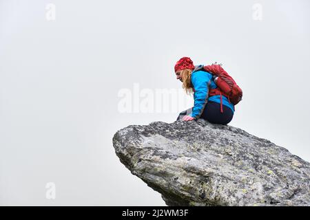 Donna escursionista con zaino rosso siede al bordo di una scogliera e guarda verso il basso, isolato dalla nebbia. Escursioni in Slovacchia alta Tatra montagne. Foto Stock