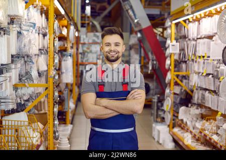 Addetto alle vendite in uniforme presso il negozio di hardware. Uomo in piedi accanto agli scaffali con strumenti, sorridente Foto Stock