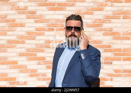 Ritratto di uomo d'affari con occhiali da sole che parlano al telefono contro muro di mattoni Foto Stock