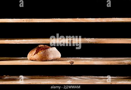 Una pagnotta di pane cotto su ripiani vuoti di legno con farina in panetteria leggera su sfondo nero durante lo scarso tempo Foto Stock