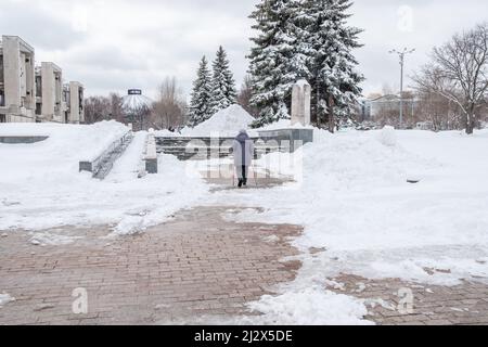 Una donna anziana con canne nelle mani cammina lungo un sentiero innevato della città. Una donna anziana allena il Nordic Walking con bastoni in città in primavera ad aprile. Foto Stock