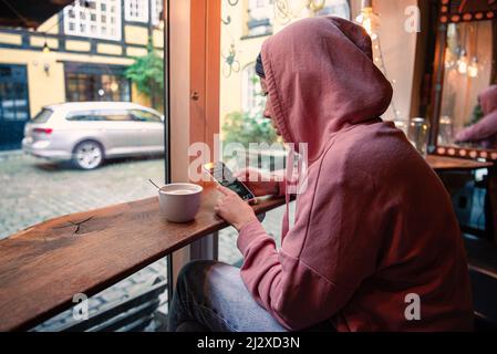 Una ragazza con capelli neri in una felpa con cappuccio e jeans guarda nel suo smartphone e beve un cappuccino caffè mentre si siede vicino alla finestra in un bar. Copenha Foto Stock