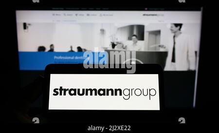 Persona che tiene il cellulare con il logo della società medica svizzera Straumann Holding AG sullo schermo di fronte al sito web aziendale. Mettere a fuoco sul display del telefono. Foto Stock