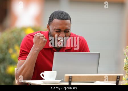 Un uomo emozionato con la pelle nera che controlla i contenuti del laptop che festeggia in una terrazza bar Foto Stock