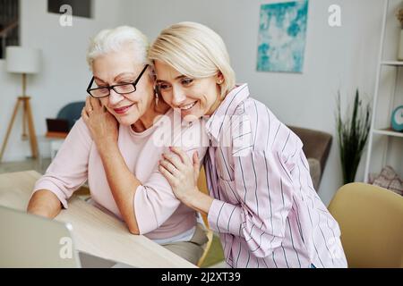Ritratto di giovane donna sorridente che abbraccia la madre anziana mentre guarda insieme lo schermo del laptop in un ambiente domestico accogliente Foto Stock