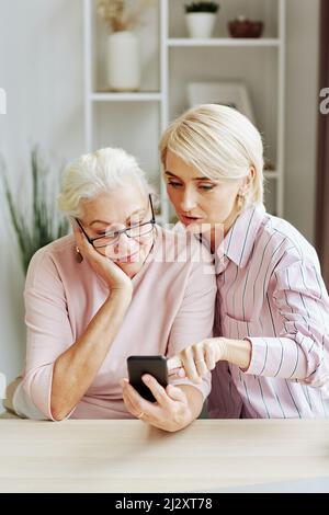 Ritratto verticale della giovane donna bionda che aiuta la madre anziana usando lo smartphone in casa accogliente Foto Stock