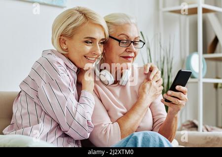 Ritratto della donna anziana moderna con smartphone a casa con una figlia sorridente che la abbraccia Foto Stock