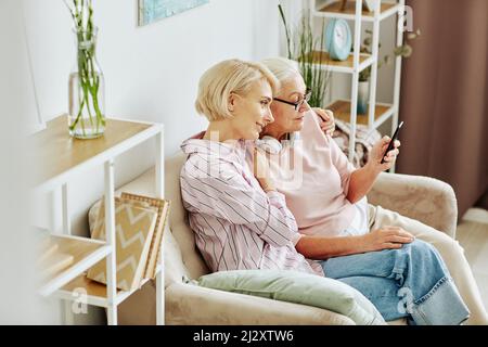 Ritratto ad angolo alto di figlia e madre adulta seduta sul divano e guardando lo schermo dello smartphone in casa accogliente Foto Stock