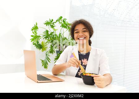 Felice African American femmina medico avendo sano pranzo seduto al tavolo, focalizzato nero donna medico consulenza online con laptop. Salute e di Foto Stock