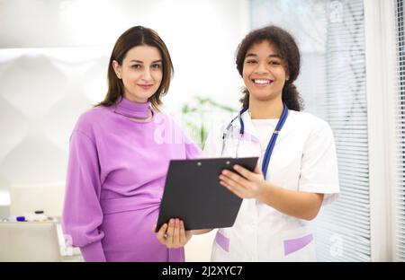 Bella giovane medico misto con paziente incinta seduta davanti al partecipante, prescrizione scritta sul modulo. Medicina e farmacia conce Foto Stock