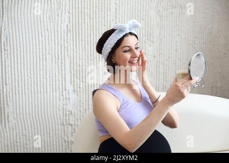 Felice bella bruna gravidanza giovane donna applicare crema al mattino guardando allo specchio circondato da pantaloni. Concetto di bellezza e assistenza sanitaria Foto Stock