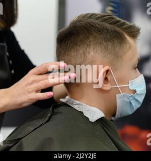 Un ragazzo adolescente ottiene un taglio di capelli in un barbiere durante una pandemia, un taglio di capelli nel salone, un cliente e un parrucchiere in maschere, un taglio di capelli del bambino con sciss Foto Stock