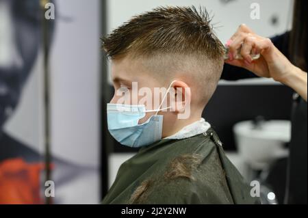 Un ragazzo adolescente ottiene un taglio di capelli in un barbiere durante una pandemia, un taglio di capelli nel salone, un cliente e un parrucchiere in maschere, un taglio di capelli del bambino con sciss Foto Stock