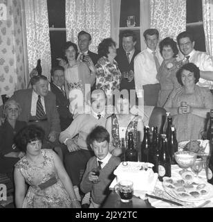 1961, storico, fidanzato partito, fidanzato coppia stting su una sedia divano insieme, circondato da amici e famiglia, Stockport, Manchester, Inghilterra, Regno Unito. Una torta celebrativa è sul tavolo come le bottiglie di birra, mentre seduto accanto al tavolo, sorella dà un'occhiata al fratello più giovane! Foto Stock