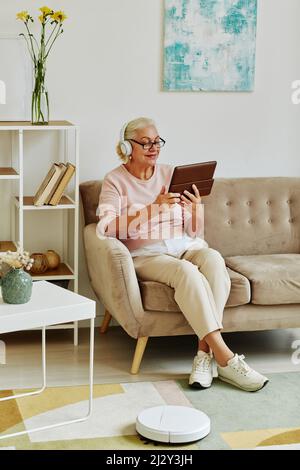 Ritratto verticale a tutta lunghezza di una donna anziana moderna utilizzando un tablet con cuffie che si trovano sul divano in un elegante ambiente domestico Foto Stock