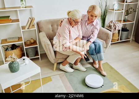Ritratto ad angolo elevato di donna anziana che utilizza l'aspirapolvere robot tramite smartphone durante la configurazione di dispositivi domestici intelligenti con l'aiuto di una figlia adulta Foto Stock