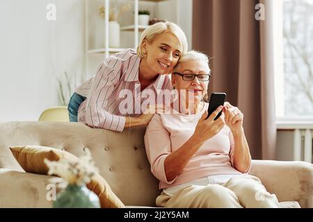 Ritratto di sorridente figlia adulta con madre usando lo smartphone insieme in casa accogliente scena Foto Stock