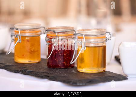 Tre pentole di conserve per toast o croissant in piccoli vasetti di vetro su un tavolo per la colazione. I vasetti contengono miele, marmellata e confettura di fragole. Foto Stock