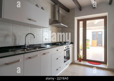 Cucina bianca con luce naturale dal cortile. Mobili bianchi in marmo nero. Soffitto con travi in legno. Piano cottura a gas ed estrattore in acciaio inox fa Foto Stock