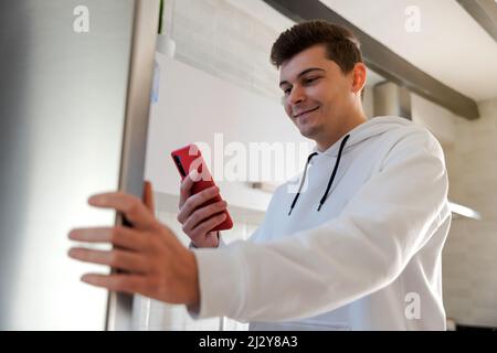 L'uomo caucasico sorride in cucina facendo la lista della spesa. Sta controllando la lista di shopping sul suo telefono mobile.he è vestito in un sudatshir bianco Foto Stock