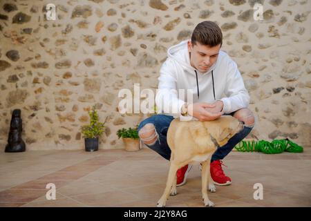 Giovane uomo caucasico sorridente mentre accarezza il suo cane nel patio all'aperto. Sfondo da parete in pietra. Felpa bianca. Spagna, casa rurale europea. Foto Stock