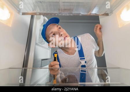 Un tecnico di sesso maschile in tute ripara un frigorifero rotto con un cacciavite. Concetto di riparazione frigorifero. Foto dall'interno del frigorifero Foto Stock