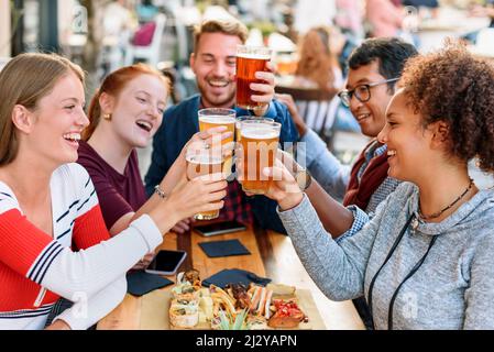 Gruppo di amici multirazziali positivi che si aggraffano bicchieri di birra mentre si siede a tavola con spuntini mentre festeggia la vacanza sulla terrazza del pub all'aperto Foto Stock