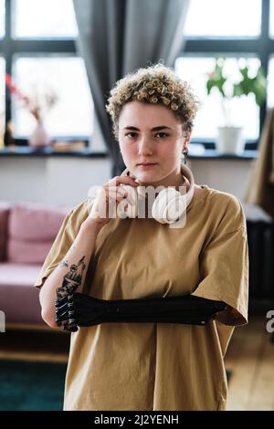 Ritratto di giovane donna con braccio protesico che ascolta musica in cuffie wireless in piedi nella stanza Foto Stock