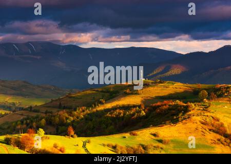 paesaggio rurale montuoso al tramonto nuvoloso. pendii boschivi con campi rurali alla luce della sera. villaggio nella valle distante dietro la collina. crinale Foto Stock