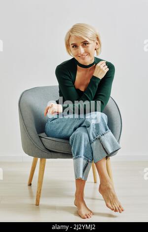 Ritratto minimo di tutta la lunghezza di elegante giovane donna seduta in poltrona e guardando la macchina fotografica in studio Foto Stock