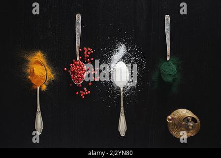 Vari tipi di spezie visualizzate su sfondo nero in cucchiai da tè. Vista dall'alto su curcuma, pepe rosso, peperoncino, peperoncino, sale e girulina Foto Stock