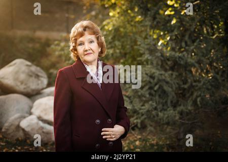 Una donna russa matura con capelli corti si erge nella foresta in un cappotto rosso bottonato. Foto Stock