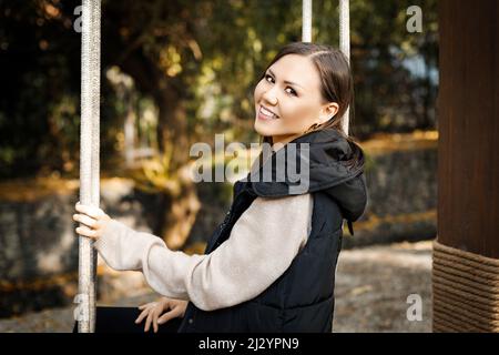 Bella giovane sorridente donna seduta su un altalena in autunno in un parco all'aperto. Foto di alta qualità Foto Stock
