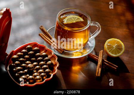 Una tazza di tè caldo appena fatto con limone, cannella e cioccolatini al sole su sfondo di legno scuro Foto Stock