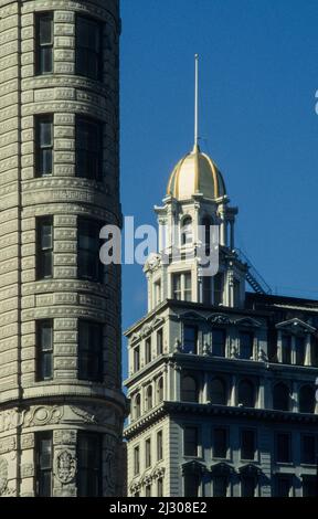 Una tranquilla mattina presto a New York City: Il famoso Flatiron Building che è stato costruito nel 1902 si trova all'incrocio tra 5th Avenue e Broadway che si incontrano qui in un angolo appuntito. Sullo sfondo la cupola in cima all'angolo-edificio 22nd ° e 5th. Foto Stock