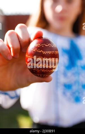 Defocus ragazza ucraina in vyshyvanka tenendo un uovo rosso colorato su sfondo naturale. Pasqua, Ucraina. Uova dipinte di mestiere. Pysanka o krashanka. Fuori Foto Stock