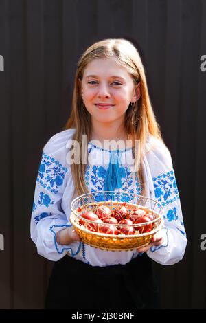 Defocus ragazza sorridente ucraina in vyshyvanka che tiene un cestino di uova rosse colorate su sfondo nero. Pasqua, Ucraina. Uova di mestiere. Raccolta di pysank Foto Stock