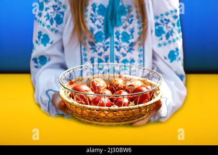 Defocus ragazza ucraina in vyshyvanka che tiene un cestino di uova rosse colorate su sfondo blu e giallo. Pasqua, bandiera Ucraina. Uova di mestiere. Raccolta di Foto Stock