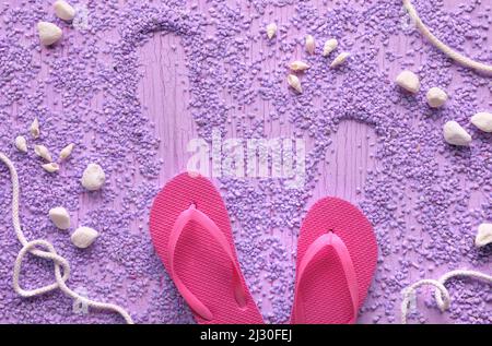Estate al mare. Infradito in gomma magenta, pantofole su ciottoli con conchiglie e ciottoli, studio concept. Sfondo viola con texture, posto per il testo. Foto Stock