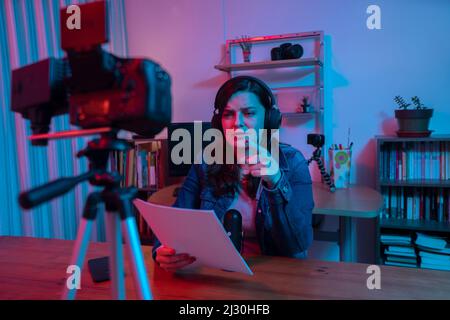 Bella donna ispanica di fronte a una videocamera che registra un blog nel suo studio con luci rosse e blu all'interno della sua casa. Streaming di un discorso su così Foto Stock