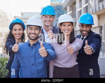 Lasciate l'edificio a noi. Sparato di un gruppo diversificato di uomini d'affari in piedi insieme e mostrando un pollice in su mentre indossa i hardhats. Foto Stock
