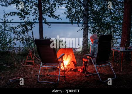 Donna seduta accanto al fuoco nella foresta sulle rive del lago Siljan a Dalarna, Svezia Foto Stock