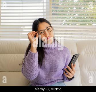 Ragazza asiatica con occhiali, cuffie e piante che lavorano da casa con un computer portatile e cellulare. Foto Stock